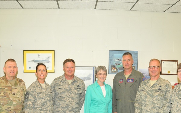 A group of seven individuals pose for a formal photo in what appears to be a military office setting. Six of the individuals are wearing military uniforms — a mix of Army combat uniforms and Air Force ABUs (Airman Battle Uniforms), along with one person in a flight suit — while one civilian woman in a teal blouse and khaki pants stands in the center. The setting suggests an official visit or meeting at a military installation.