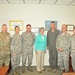 A group of seven individuals pose for a formal photo in what appears to be a military office setting. Six of the individuals are wearing military uniforms — a mix of Army combat uniforms and Air Force ABUs (Airman Battle Uniforms), along with one person in a flight suit — while one civilian woman in a teal blouse and khaki pants stands in the center. The setting suggests an official visit or meeting at a military installation.