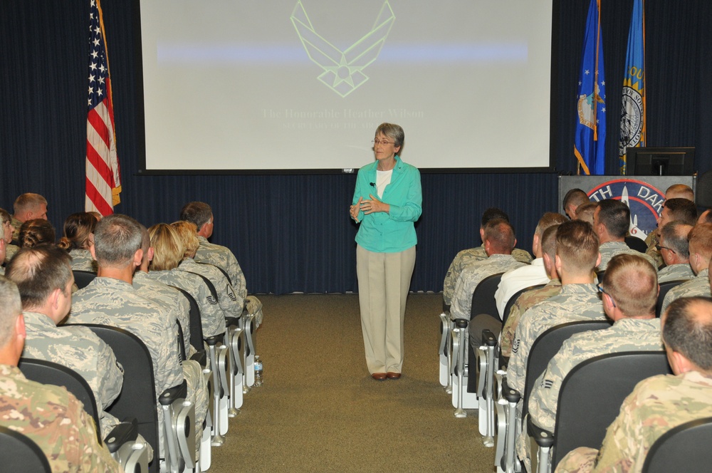 A civilian woman in a teal blazer and khaki pants stands at the front of a briefing room addressing a large audience of uniformed military personnel seated in rows. The projection screen behind her displays the U.S. Air Force symbol and text reading 'The Honorable Heather Wilson' along with additional text. The setting appears to be a formal military briefing or town hall event at a North Dakota Air National Guard or Air Force installation.