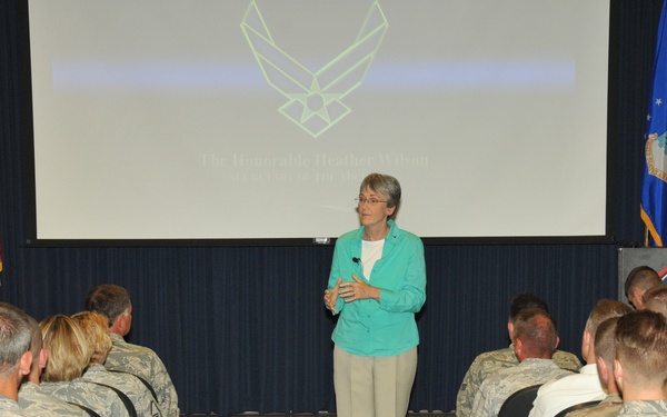 A civilian woman in a teal blazer and khaki pants stands at the front of a briefing room addressing a large audience of uniformed military personnel seated in rows. The projection screen behind her displays the U.S. Air Force symbol and text reading 'The Honorable Heather Wilson' along with additional text. The setting appears to be a formal military briefing or town hall event at a North Dakota Air National Guard or Air Force installation.