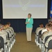 A civilian woman in a teal blazer and khaki pants stands at the front of a briefing room addressing a large audience of uniformed military personnel seated in rows. The projection screen behind her displays the U.S. Air Force symbol and text reading 'The Honorable Heather Wilson' along with additional text. The setting appears to be a formal military briefing or town hall event at a North Dakota Air National Guard or Air Force installation.