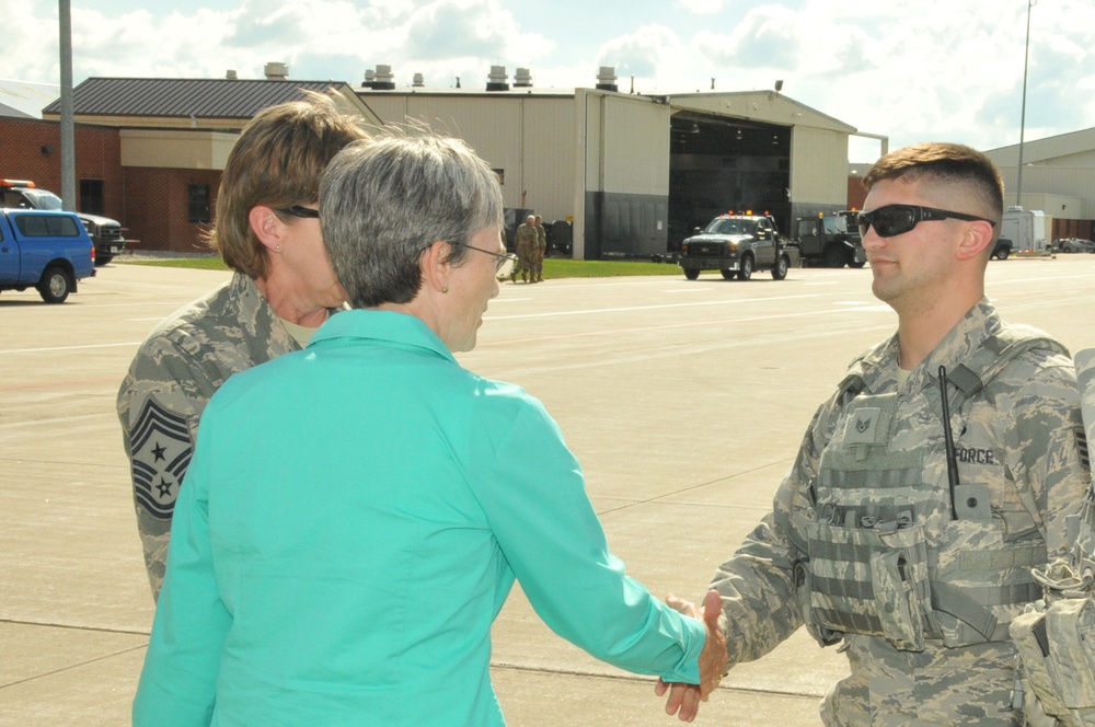 A civilian woman in a teal blazer is shaking hands with a US Air Force airman in ABU (Airman Battle Uniform) on what appears to be a military flight line or tarmac. A senior enlisted Air Force member wearing a Chief Master Sergeant rank insignia is visible in the background. The setting appears to be an official visit or greeting ceremony at a military air base.