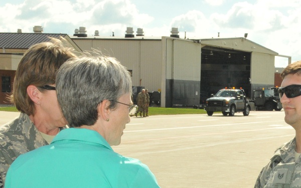 A civilian woman in a teal blazer is shaking hands with a US Air Force airman in ABU (Airman Battle Uniform) on what appears to be a military flight line or tarmac. A senior enlisted Air Force member wearing a Chief Master Sergeant rank insignia is visible in the background. The setting appears to be an official visit or greeting ceremony at a military air base.