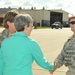 A civilian woman in a teal blazer is shaking hands with a US Air Force airman in ABU (Airman Battle Uniform) on what appears to be a military flight line or tarmac. A senior enlisted Air Force member wearing a Chief Master Sergeant rank insignia is visible in the background. The setting appears to be an official visit or greeting ceremony at a military air base.