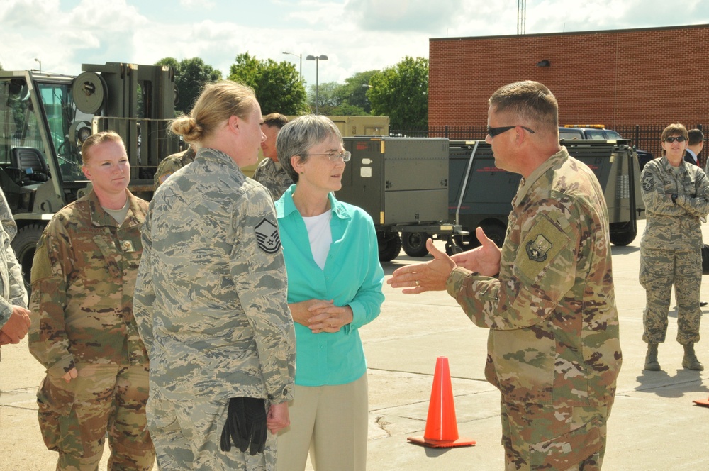 A civilian woman in a teal jacket is engaged in conversation with military personnel in an outdoor military installation setting. The group includes both Air Force members in ABU (Airman Battle Uniform) pattern and soldiers in OCP (Operational Camouflage Pattern) uniforms. The setting appears to be a flight line or equipment staging area with military vehicles and equipment visible in the background.