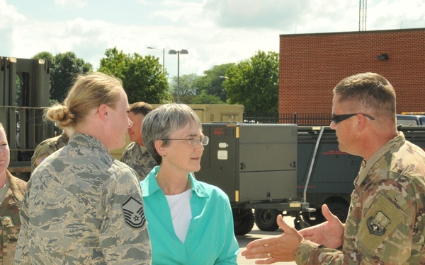 A civilian woman in a teal jacket is engaged in conversation with military personnel in an outdoor military installation setting. The group includes both Air Force members in ABU (Airman Battle Uniform) pattern and soldiers in OCP (Operational Camouflage Pattern) uniforms. The setting appears to be a flight line or equipment staging area with military vehicles and equipment visible in the background.