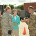 A civilian woman in a teal jacket is engaged in conversation with military personnel in an outdoor military installation setting. The group includes both Air Force members in ABU (Airman Battle Uniform) pattern and soldiers in OCP (Operational Camouflage Pattern) uniforms. The setting appears to be a flight line or equipment staging area with military vehicles and equipment visible in the background.