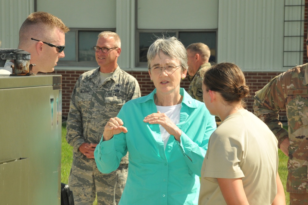 A civilian woman in a teal blouse is engaged in conversation with military personnel outdoors near a building. She appears to be speaking with a female soldier in tan/multicam uniform while other Air Force and Army personnel stand nearby. The setting appears to be a military installation or base.