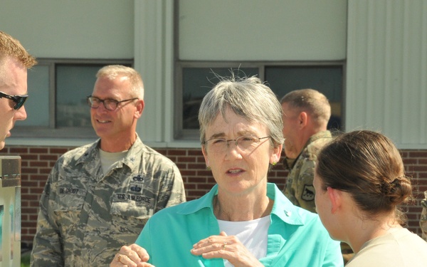 A civilian woman in a teal blouse is engaged in conversation with military personnel outdoors near a building. She appears to be speaking with a female soldier in tan/multicam uniform while other Air Force and Army personnel stand nearby. The setting appears to be a military installation or base.