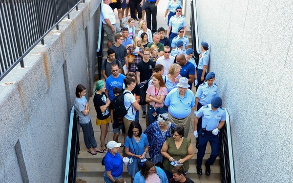 An aerial/overhead view of a large crowd of civilians and military personnel in light blue uniforms moving through a narrow outdoor corridor or stairwell passage between two concrete walls. The group appears to be attending a guided tour or organized event, with military escorts visible throughout the crowd. The image is taken from a significant height looking straight down at the crowd below.