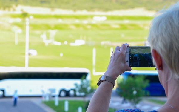 A woman with short blonde hair is photographing a scene below from an elevated vantage point using a smartphone. The scene below shows a large white coach bus parked near a road, with a long queue of people lined up along a railing. The setting appears to be a stadium or sports facility with green fields visible in the background.