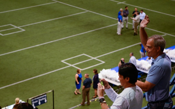 The image shows spectators standing in the lower seating area of an indoor stadium or arena, overlooking a green artificial turf field. Several people are taking photos with their phones while others mill about on the field below. The event appears to be a pre-game, open practice, or special access event given the informal atmosphere and table setups on the field.