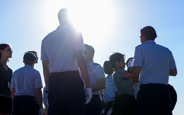 A group of individuals, several wearing white military-style uniforms with dark trousers, are gathered outdoors on a paved surface in bright sunlight. The strong backlighting from a low sun creates silhouettes and lens flare, making some figures difficult to distinguish. At least one civilian in athletic wear (dark shirt and shorts) is visible among the uniformed personnel.