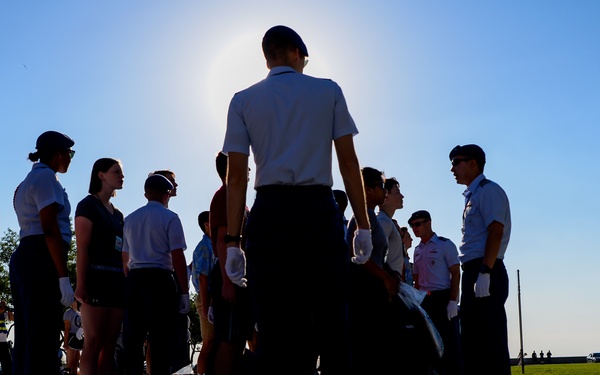 A group of individuals in military-style uniforms and civilian clothing are gathered outdoors on a paved surface, appearing to be engaged in a drill or training exercise. The image is taken against a bright sun, creating strong silhouettes and dramatic shadow patterns on the ground. A photographer is visible in the background documenting the event.