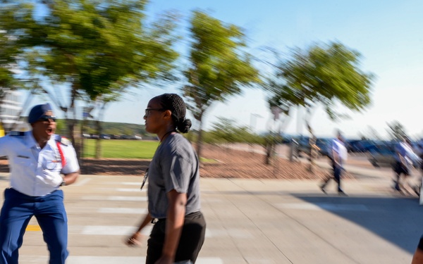 A uniformed military or ROTC cadre member appears to be directing or shouting at two young individuals carrying bags as they cross a marked crosswalk. The scene appears to take place on a military academy or training facility campus, with modern buildings and trees visible in the background. The motion blur effect suggests dynamic movement and urgency in the scene.