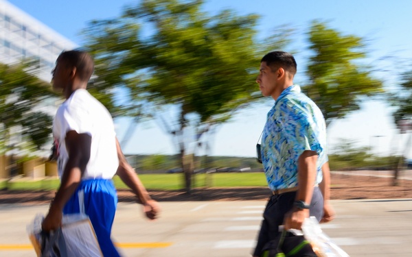 Two young men are walking briskly across a pedestrian crosswalk, each carrying large duffel bags or backpacks. The image features intentional motion blur, suggesting fast movement or a panning camera technique. The setting appears to be an urban or campus environment with trees and a modern building visible in the background.