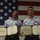 Two uniformed military personnel are posing in front of a large American flag, each holding official award certificates. The documents visible are 'Air Medal' citations issued by The United States of America. Both individuals are wearing light blue uniforms with ribbons and insignia, consistent with U.S. Air Force or Coast Guard dress uniforms.
