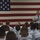 A senior military officer addresses a large assembly of uniformed personnel in what appears to be a hangar or large indoor facility. The speaker stands at the front holding a microphone, with a large American flag displayed prominently on the wall behind him. The audience, dressed in light blue uniforms consistent with U.S. Coast Guard or Air Force dress uniforms, is seated and facing the speaker.