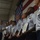 A military ceremony is taking place indoors with uniformed personnel standing in a line at attention. An officer on the left appears to be pinning medals or insignia on the personnel, suggesting an awards or promotion ceremony. A large American flag is displayed prominently on the wall in the background.