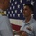A military award ceremony is taking place in front of a large American flag. A senior officer with gold captain's insignia on his collar is pinning a medal or award onto a junior service member wearing a light blue uniform with aviator wings and service ribbons. The recipient stands at attention with a composed expression.