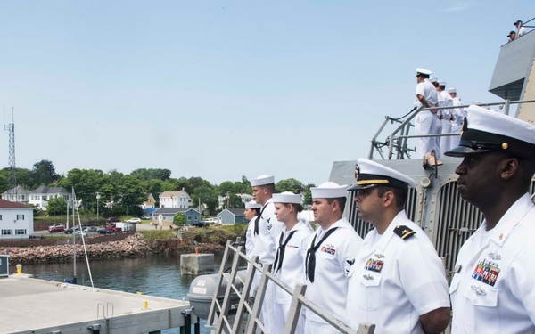 A group of U.S. Navy personnel in white dress uniforms are standing at attention on the deck and gangway of a naval vessel, likely during a formal arrival or departure ceremony at a naval installation. Officers and enlisted sailors are arranged in a line facing the pier, with additional crew members visible on upper deck levels. The setting appears to be a coastal naval base with buildings, trees, and a concrete pier visible in the background.