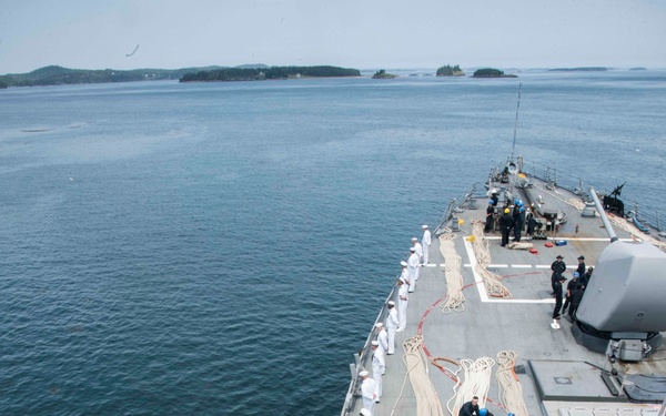 An aerial view of a naval warship deck showing sailors in white dress uniforms lined along the port side rail, consistent with a 'manning the rails' ceremonial formation during a port arrival or departure. Large coiled mooring lines are laid out on the forward deck, and several crew members in working uniforms are managing deck equipment. The vessel is navigating calm waters with tree-covered islands visible in the background.
