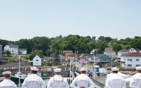 Eight U.S. Navy sailors in dress white uniforms stand at parade rest along the rail of a naval vessel, facing a small coastal town harbor. The scene appears to be a port visit or homecoming event, with a crowd of civilians visible on the dock below. An American flag is visible in the background near the waterfront buildings.