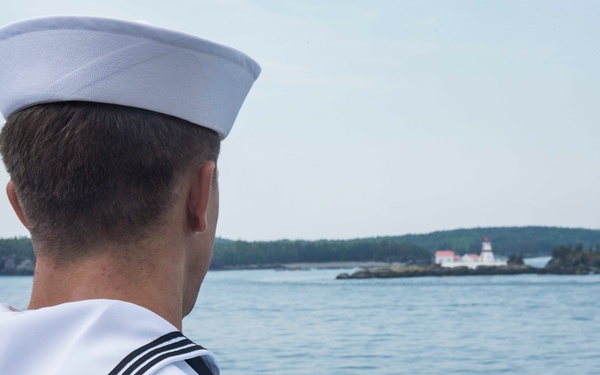 A U.S. Navy sailor in dress white uniform is seen from behind, standing aboard a vessel and looking out toward a coastal lighthouse on a wooded island. The sailor's uniform patch identifies the ship as the USS McFaul. The scene appears to be taken during calm daylight conditions on open coastal waters.