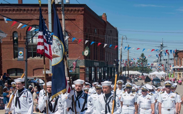 A U.S. Navy color guard and personnel are marching in a public parade down a main street of a small town. They are carrying the American flag and Navy flag while holding a banner reading 'DDG-74' with the words 'Courage - Honor - Sacrifice.' Spectators line the sidewalks and the street is decorated with red, white, and blue bunting.