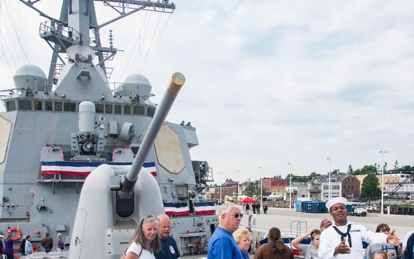 A group of civilians is touring the deck of a U.S. Navy warship, likely during a public open-ship event. A Navy sailor in dress whites is serving as a guide, gesturing and explaining features of the vessel to the visitors. The ship appears to be docked at a harbor with a small coastal town visible in the background.