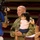 A U.S. Army soldier in camouflage uniform is holding a young infant while being filmed by a television camera crew. The soldier is smiling broadly, suggesting a joyful occasion, likely a military homecoming or recognition event. The setting appears to be an indoor arena or gymnasium with stadium seating visible in the background.