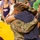 A U.S. Army soldier in OCP (Operational Camouflage Pattern) uniform is sharing an emotional embrace with a civilian, both appearing visibly moved. The soldier displays a U.S. flag patch and a unit patch on the left sleeve, consistent with an active duty Army uniform. The scene appears to take place in a stadium or arena setting with spectators visible in the background.