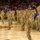 A military formation ceremony is taking place on the hardwood court of what appears to be Arizona State University's arena (Wells Fargo Arena), identifiable by the maroon and gold color scheme and ASU logo on the floor. Dozens of soldiers in Army Combat Uniforms (ACUs/OCP camouflage pattern) are standing at attention in organized ranks, with several guidon bearers holding blue unit flags. A large crowd of civilian spectators fills the arena seating in the background.