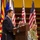 A civilian man in a dark suit is speaking at a wooden podium with a microphone at what appears to be an official military or government ceremony. Multiple flags are visible in the background, including American flags and what appears to be the Arizona state flag. A military officer in camouflage uniform is seated in the background.