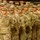 A large formation of U.S. Army soldiers standing at attention in what appears to be an indoor facility. The soldiers are wearing Operational Camouflage Pattern (OCP) uniforms with unit patches and name tapes visible. The formation suggests a formal military ceremony, briefing, or muster event.