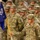 A formation of U.S. Army soldiers in Operational Camouflage Pattern (OCP) uniforms stands at attention during what appears to be a formal military ceremony. A soldier in the foreground, identified by nametape as 'GUNN,' holds a blue guidon flag bearing the number '158.' The scene suggests a unit formation, possibly a deployment ceremony, activation, or awards event.