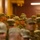 A large group of military personnel in matching camouflage uniforms and patrol caps are assembled indoors, appearing to attend a briefing or formation. The soldiers are wearing what appears to be the US Army Operational Camouflage Pattern (OCP) uniforms. The setting appears to be a military facility or auditorium with warm ambient lighting.