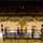 A military ceremony is taking place inside what appears to be Arizona State University's Wells Fargo Arena, identifiable by the maroon and gold court markings and 'Sun Devil' logo. A large formation of soldiers in camouflage uniforms is assembled on the basketball court, facing a raised stage with flags including the US flag, Arizona state flag, and military branch flags. The arena has spectators in the stands observing the formal military event.