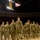 A group of U.S. Army soldiers in OCP (Operational Camouflage Pattern) uniforms are marching in formation on a basketball court inside an indoor arena. The venue displays a retired jersey numbered 13 with the name 'Harden' and an American flag hanging from the rafters, consistent with Arizona State University's Wells Fargo Arena. The ceremony appears to be a military deployment or mobilization event, evidenced by the formal formation and presence of state and national flags.