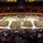A large military ceremony is taking place inside an indoor sports arena, with hundreds of soldiers in camouflage uniforms assembled in formation on the basketball court. The arena appears to be Arizona State University's Wells Fargo Arena (now Desert Financial Arena), identifiable by the maroon and gold color scheme and the ASU pitchfork logo on the court. A large civilian audience fills the lower seating sections, suggesting this is a deployment or homecoming ceremony.