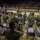 A military band in U.S. Army camouflage uniforms performs on the floor of a large indoor arena, facing a formation of soldiers standing at attention on the court. The arena appears to be filled with spectators in the stands, and American flags along with other military colors are visible near the formation. This appears to be a formal military ceremony, likely a deployment sendoff or homecoming event.