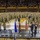 A large military ceremony is taking place inside an indoor sports arena or gymnasium, with hundreds of soldiers in camouflage uniforms assembled on the court floor. A civilian speaker stands at a podium on a raised stage in the foreground, with multiple flags displayed including the American flag. The arena stands are packed with civilian spectators observing the formal military event.