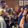 An NBC news crew is conducting an interview at what appears to be a military homecoming or deployment event. A soldier in camouflage uniform stands with a woman holding a baby while an NBC reporter holds a microphone toward them. A videographer captures the moment on a professional camera in the foreground.