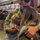 A soldier in camouflage military uniform embraces two females — appearing to be a woman and a young girl — in an emotional reunion. The setting appears to be an indoor venue, likely a gymnasium or arena, with a banner reading 'Arizona State University Sports Hall of Fame' visible in the background. The scene depicts a military homecoming event with multiple other people visible in the background.