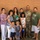 A group photo of what appears to be a multi-generational family posing together in front of a brick wall. A U.S. Army soldier in camouflage uniform (OCP/MultiCam pattern) stands at the center-right of the group, surrounded by family members including adults, children, and elderly individuals. The occasion appears to be a military family reunion or send-off event.