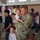 A military homecoming or deployment ceremony is taking place inside Wells Fargo Arena at Arizona State University (ASU). U.S. Army soldiers in OCP (Operational Camouflage Pattern) uniforms are reuniting with their families, including young children. The central figure is a soldier holding a toddler while a young boy stands nearby.