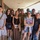 A group of approximately ten people pose together for a photo under a covered outdoor walkway or corridor. The group includes one individual in U.S. Army military camouflage uniform and a mix of civilians of varying ages, suggesting a family or group gathering. The setting appears to be a public venue or facility in a warm, sunny environment.