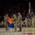 A military ceremony is taking place inside what appears to be the Wells Fargo Arena at Arizona State University (ASU), as indicated by the 'GiveTo.ASU.edu' and 'Campaign ASU 2020' banners visible in the background. Soldiers in Army Combat Uniforms (ACU) are participating in a formal flag ceremony, prominently displaying the Arizona State Flag alongside a military unit guidon. A large crowd of spectators fills the arena seating in the background.
