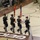 A military color guard of four soldiers in dress uniforms is marching across an indoor basketball court during a formal ceremony. The soldiers are carrying the United States flag and what appears to be the Arizona state flag, with two armed rifle bearers flanking the flag carriers. The event appears to be taking place in a sports arena with spectators and military personnel lining the sides.