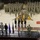 A military ceremony is taking place inside a basketball arena, identifiable by the Wells Fargo Arena court markings with a maroon and gold logo. Hundreds of soldiers in camouflage uniforms are standing in formation on the court floor while a color guard in dress uniforms presents flags on a raised stage. Dignitaries and officials stand on the stage platform observing the ceremony.
